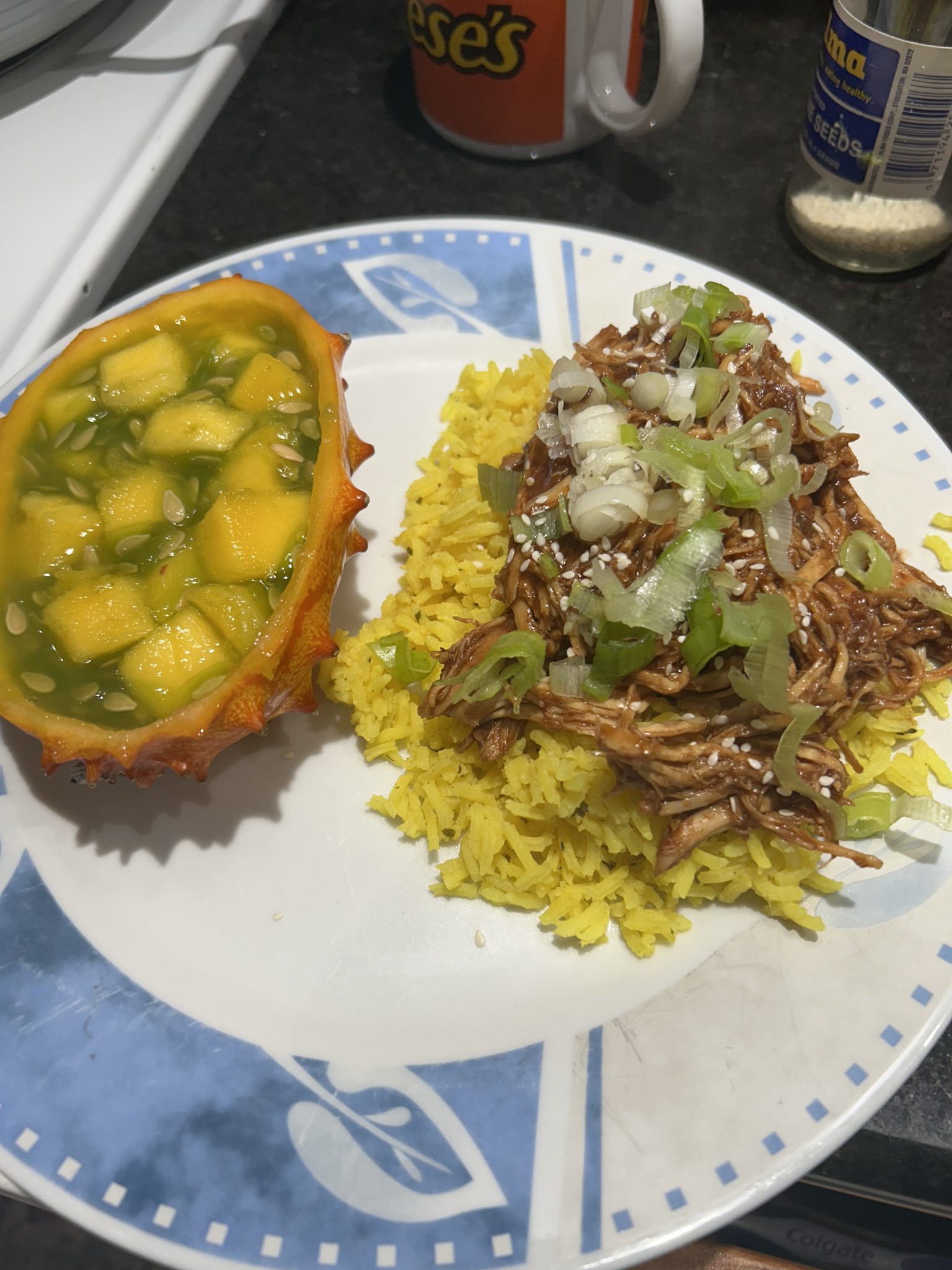 A plate with yellow rice topped with shredded chicken, green onions, and sesame seeds, served beside half a horned melon filled with yellow fruit pieces. A mug and spice jar are in the background.