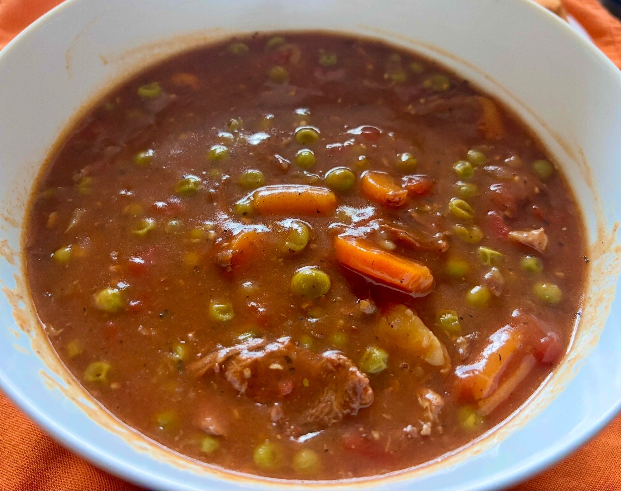 A bowl of thick, hearty stew filled with chunks of carrot, green peas, pieces of meat, and a rich, brown broth. The bowl is white and sits on an orange surface.