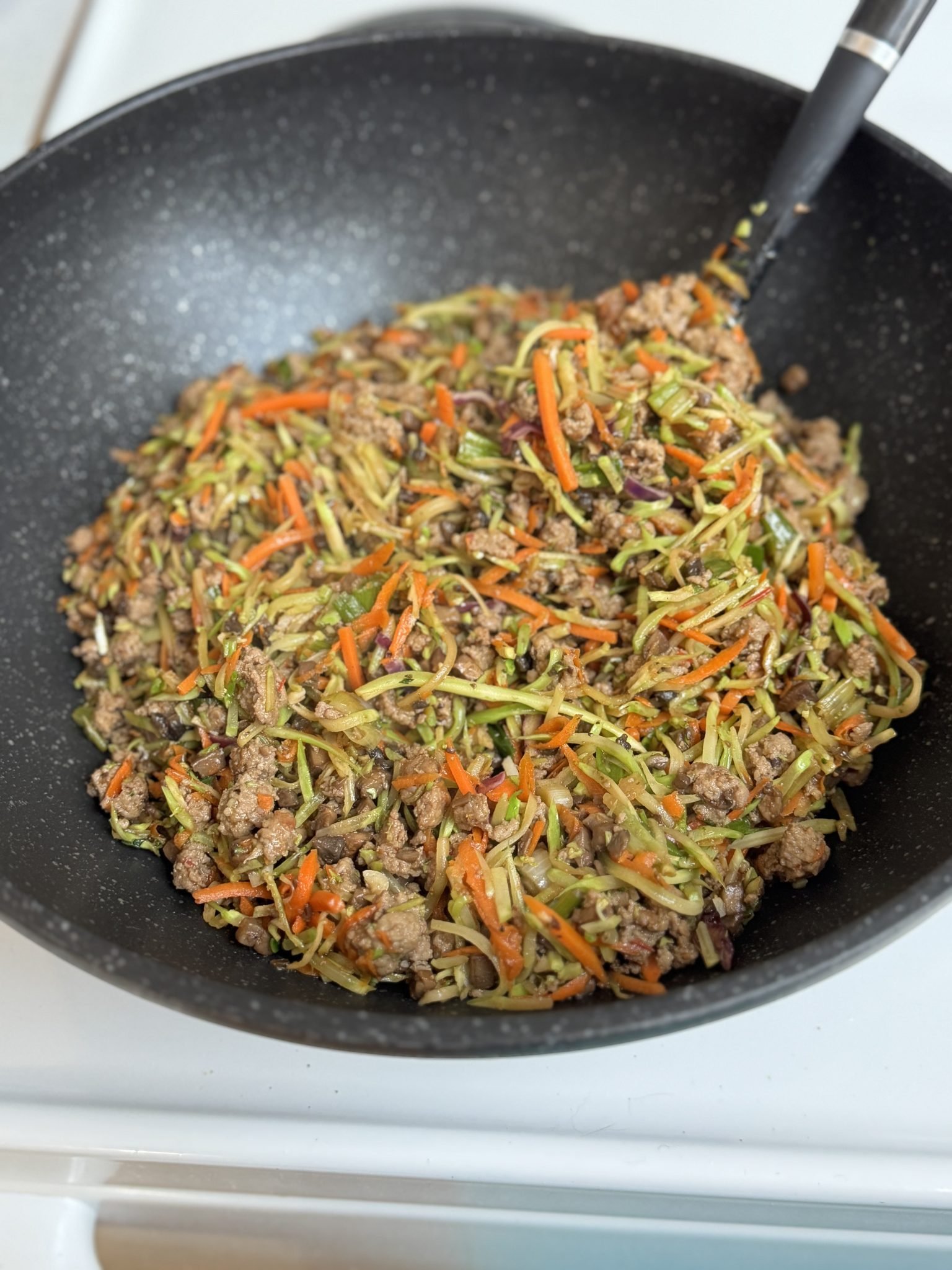 Ground meat stir fry with shredded carrots, broccoli, and cabbage in a black skillet, mixed with a spatula.