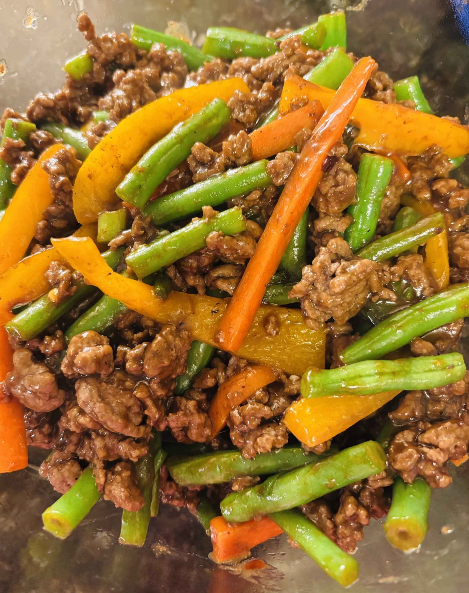 A close-up of a stir-fry dish featuring green beans, orange bell peppers, carrot strips, and ground beef coated in a savory sauce.