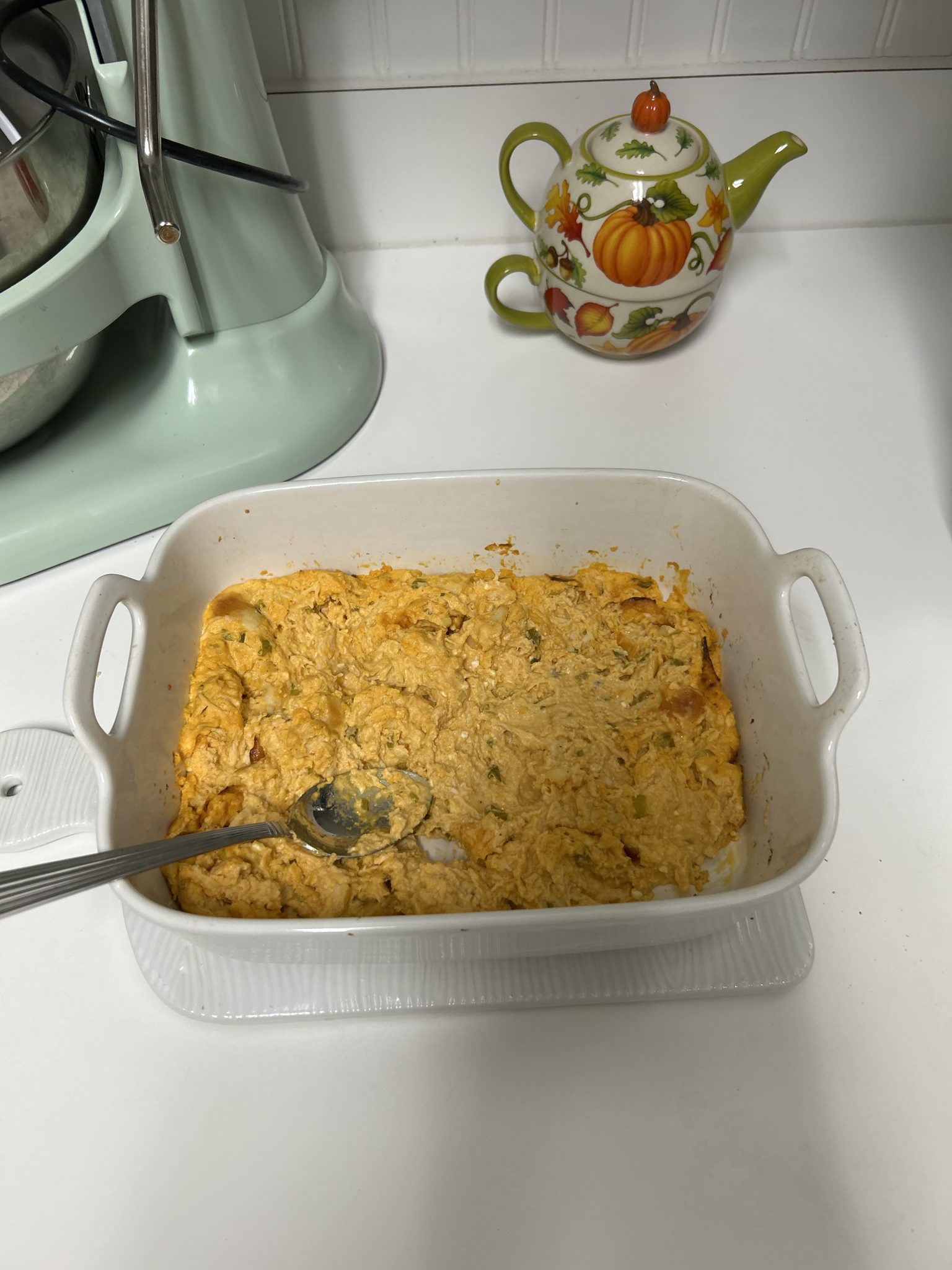 A white casserole dish with baked dip and a spoon, next to a green mixer and a pumpkin-themed teapot on the counter.