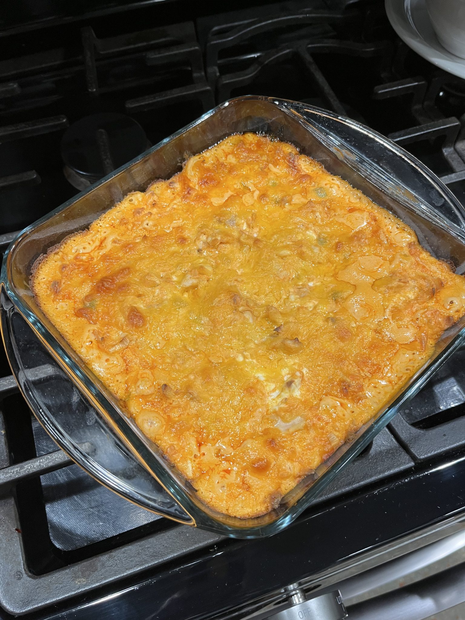 A square glass baking dish filled with a golden-brown baked delight, possibly a casserole or gratin, sits on a black stovetop. The crispy, bubbly top suggests it might be a freshly baked healthy buffalo chicken dip.