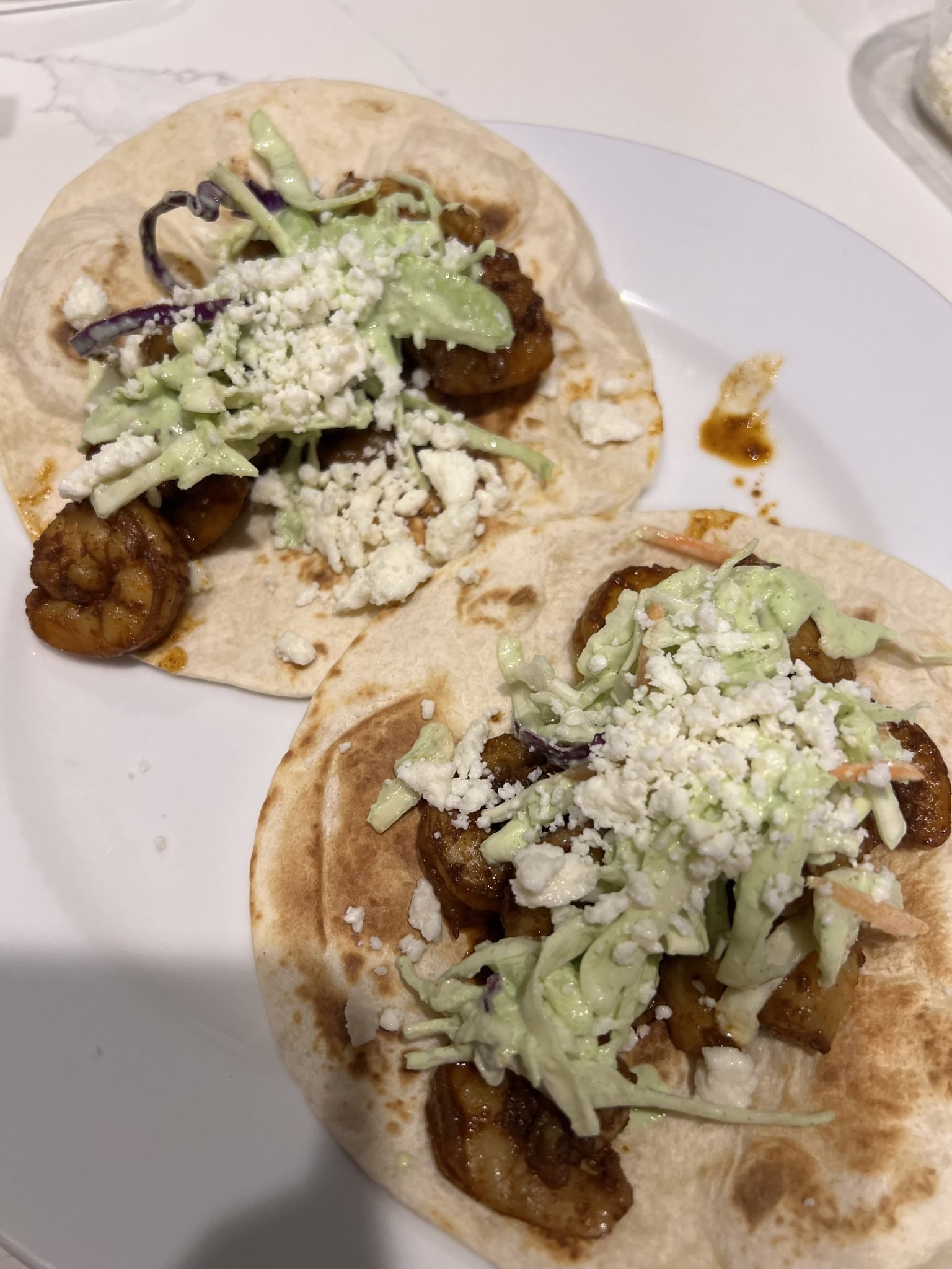 Two soft tacos on a white plate, each filled with seasoned shrimp, shredded slaw, avocado or green crema, and crumbled white cheese. The tacos are on a white surface.
