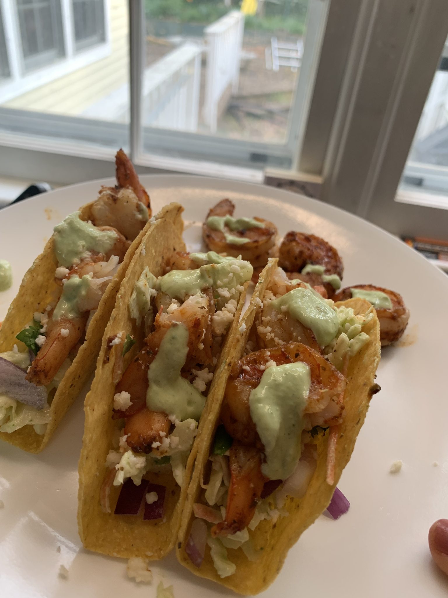 Three shrimp tacos in crunchy shells topped with creamy green sauce, crumbled cheese, lettuce, and red onion sit on a white plate near a window. Two extra shrimp are on the side of the plate.