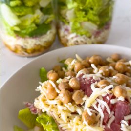 A fresh salad in a bowl topped with chickpeas, shredded cheese, and slices of salami, with two jars of layered salad containing lettuce, cheese, and other ingredients in the background.