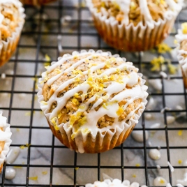Lemon poppy seed muffins topped with white icing and lemon zest, arranged on a cooling rack. The muffins are in paper liners, and some icing has dripped onto the rack below.