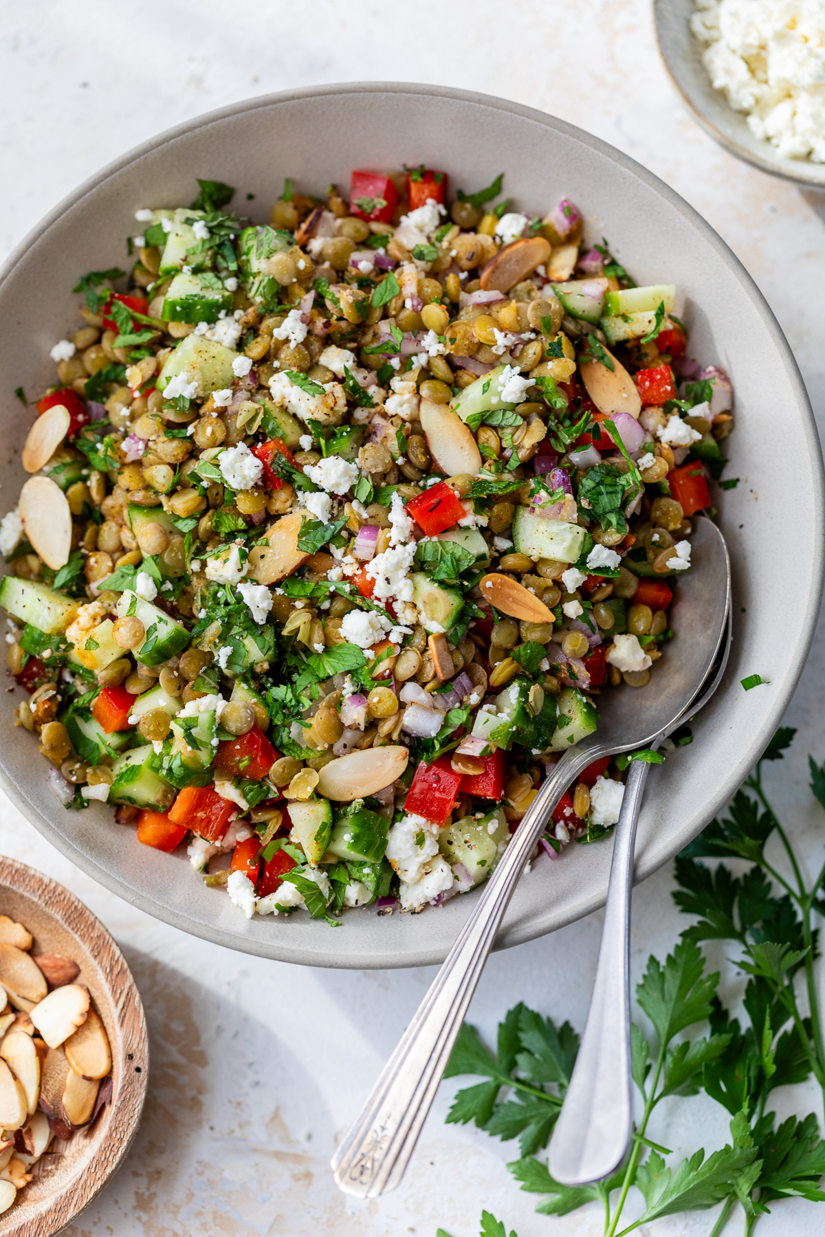A bowl of colorful lentil salad with chopped cucumber, red bell pepper, red onion, parsley, sliced almonds, and crumbled feta cheese, with two spoons in the bowl and a bunch of parsley and a bowl of almonds nearby.