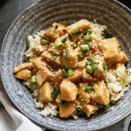 A bowl of rice topped with slow cooker orange chicken, garnished with chopped green onions and sesame seeds. The dish is served in a gray ceramic bowl on a dark surface.