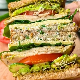 A close-up of a hand holding a thick sandwich filled with layers of tomatoes, lettuce, sprouts, avocado, cheese, and whole grain bread, with visible seeds and a colorful, fresh appearance.