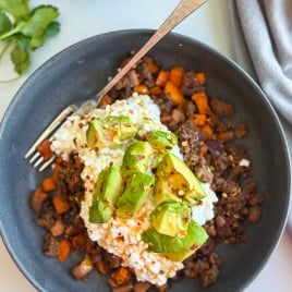 A ground beef bowl with ground beef, diced sweet potatoes, and onions, topped with cottage cheese, avocado chunks, and red pepper flakes. A silver fork rests in the bowl. Avocado, cilantro, and a gray napkin are nearby.