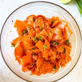A glass bowl filled with ribbon-like raw carrot salad, garnished with sesame seeds and chopped green onions. A lime slice and green onion are visible in the background.